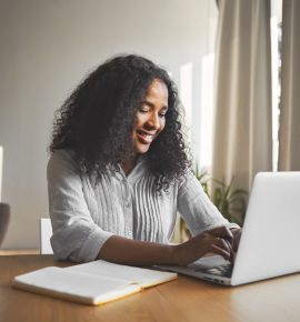 Gorgeous positive young dark skinned female blogger keyboarding on generic laptop, smiling, being inspired while creating new content for her travel blog, sitting at desk with diary and mug