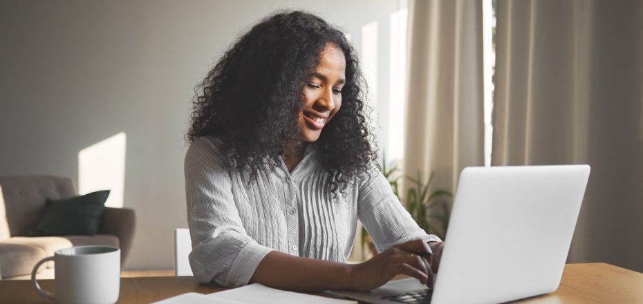 Gorgeous positive young dark skinned female blogger keyboarding on generic laptop, smiling, being inspired while creating new content for her travel blog, sitting at desk with diary and mug