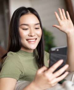 Young asian woman video calling on mobile phone, waving hand at smartphone camera, chatting on cellphone from home.